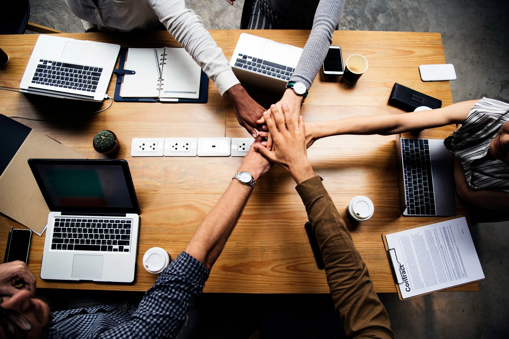 Top-down view of a business team stacking hands together in unity over a wooden table with laptops and documents